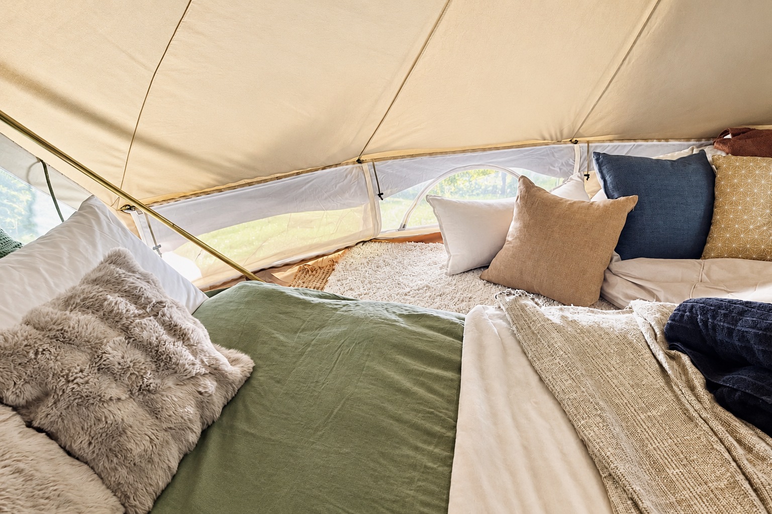 Glamping bed with white linen, cushions and layered throws inside a canvas tent.