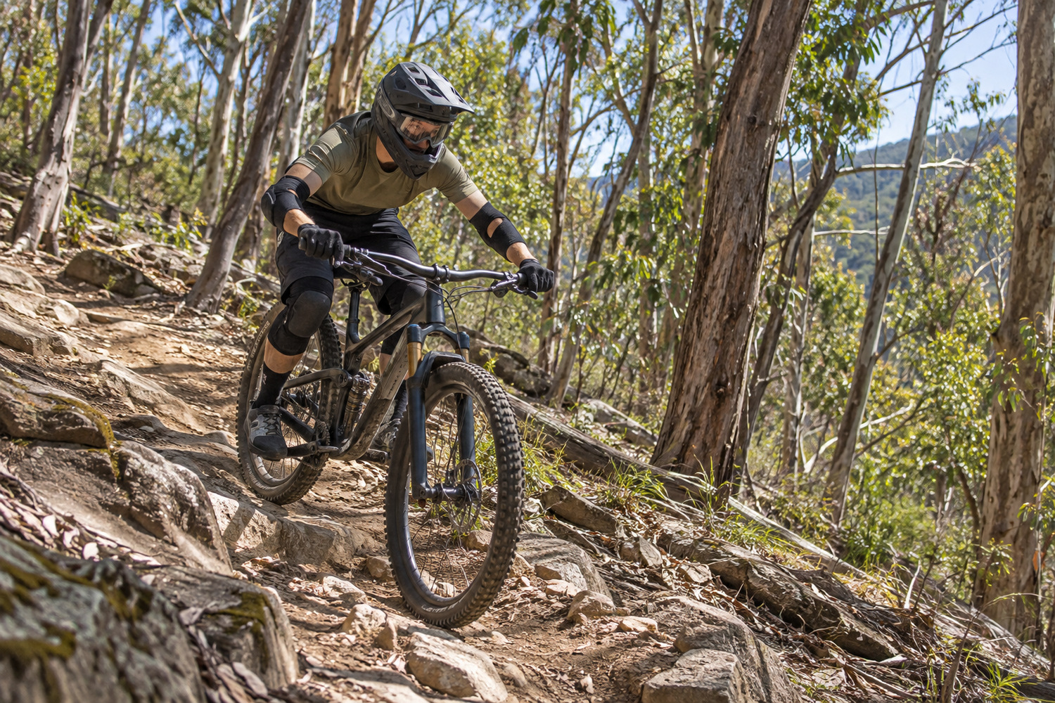 Mountain biker in a full-face helmet and protective pads descending a rocky eucalyptus forest trail.